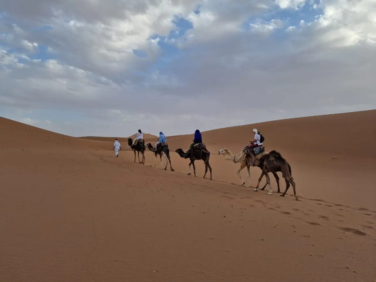 A caravan of four camels with riders and a walking guide trekking across vast desert sand dunes under a cloudy sky.