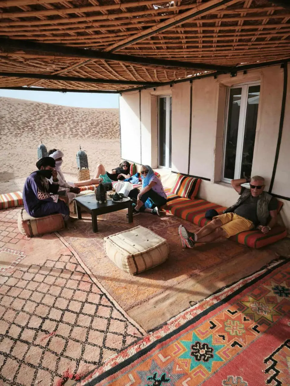 Tourists and guides relaxing on Moroccan rugs in a shaded desert camp with sand dunes in the background.