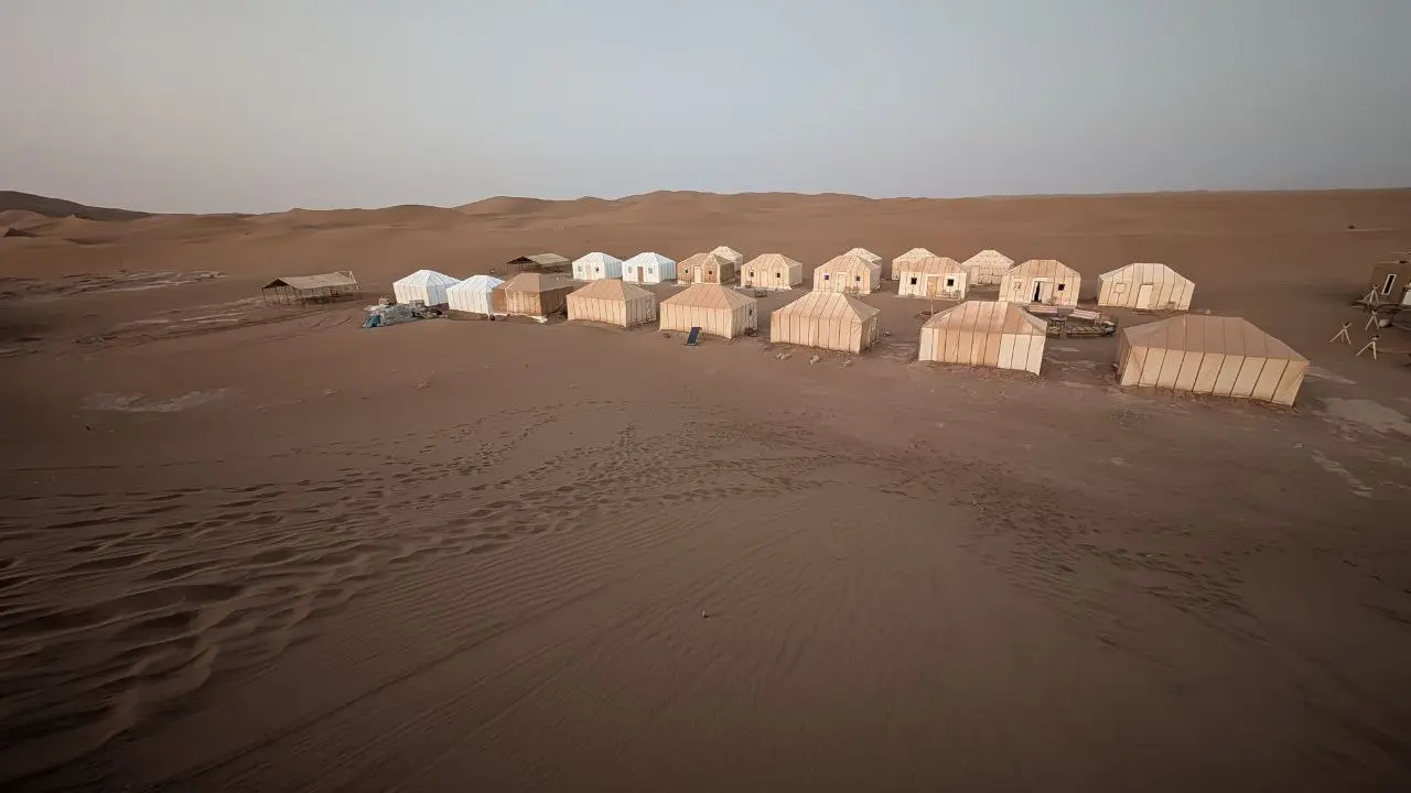A cluster of beige and white tents arranged in a desert camp surrounded by vast sand dunes.