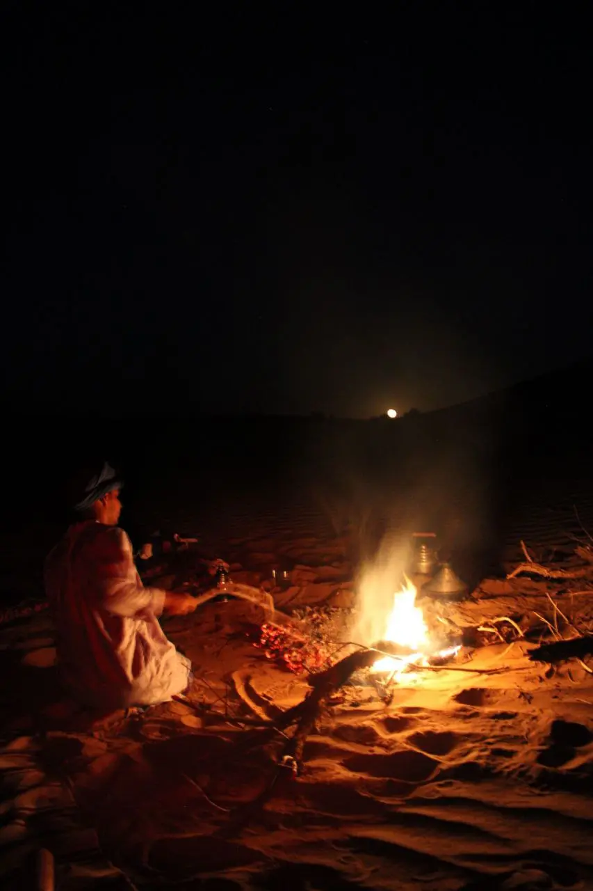 A man in traditional white robes sits by a campfire in the desert at night under a full moon.