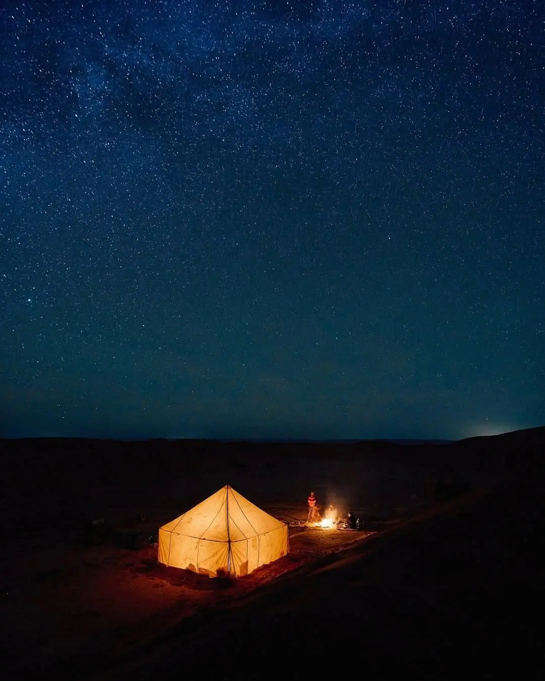 A glowing tent and campfire under a sky full of stars in a dark desert landscape.