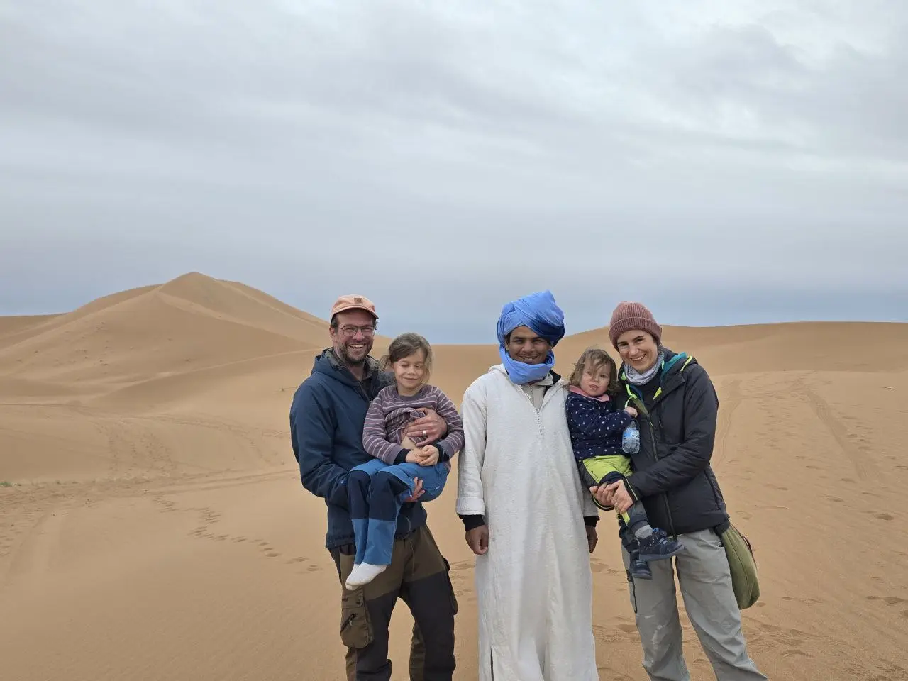 A family with two children poses with a local guide in a desert with sand dunes under a cloudy sky.