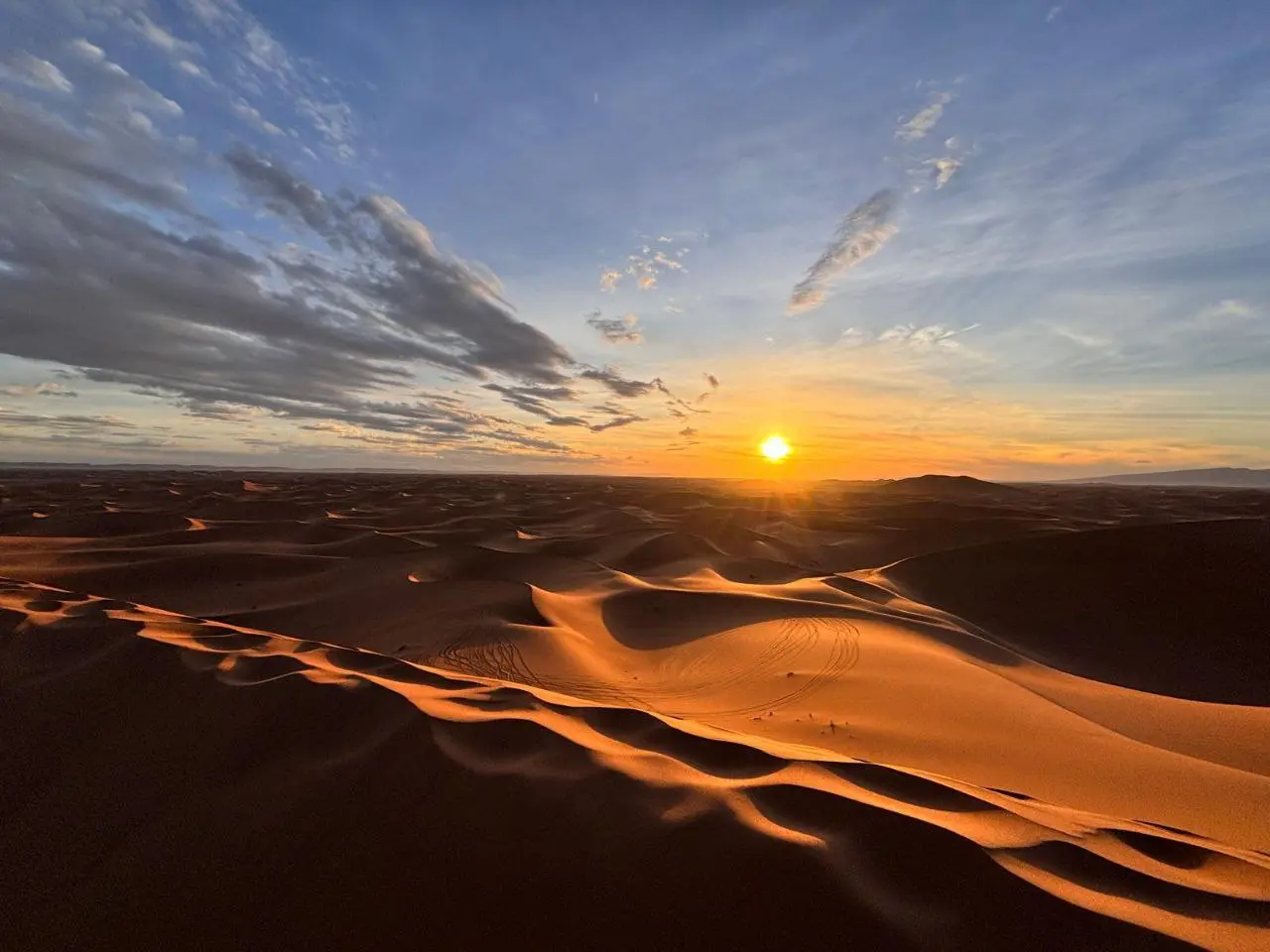 A golden sunset over vast, rolling sand dunes in a desert landscape under a streaked blue sky.