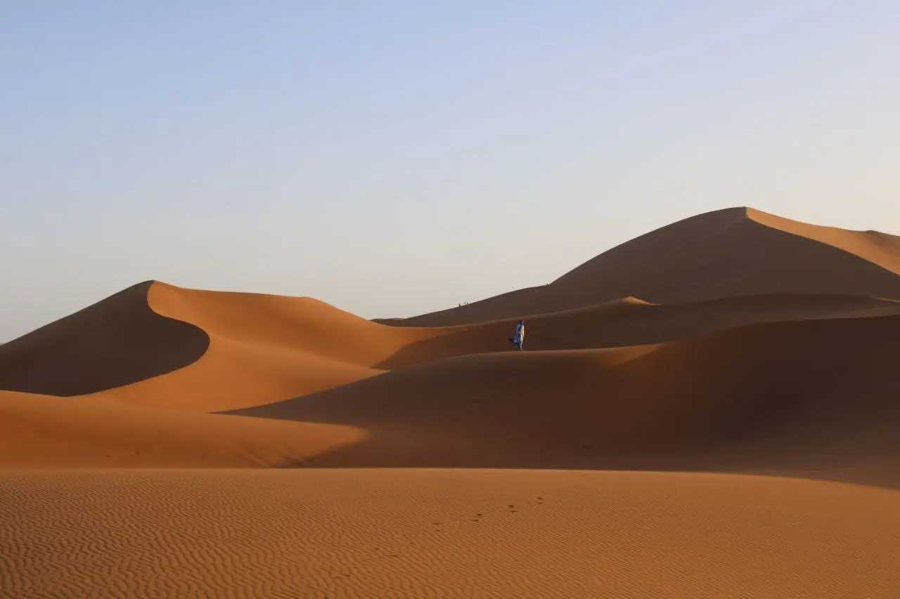 A lone traveler walking on a vast, golden sand dune in a desert under a clear sky.