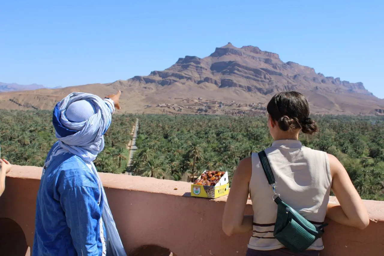 A man in traditional blue clothing points towards a mountain over a lush palm oasis for a female tourist.