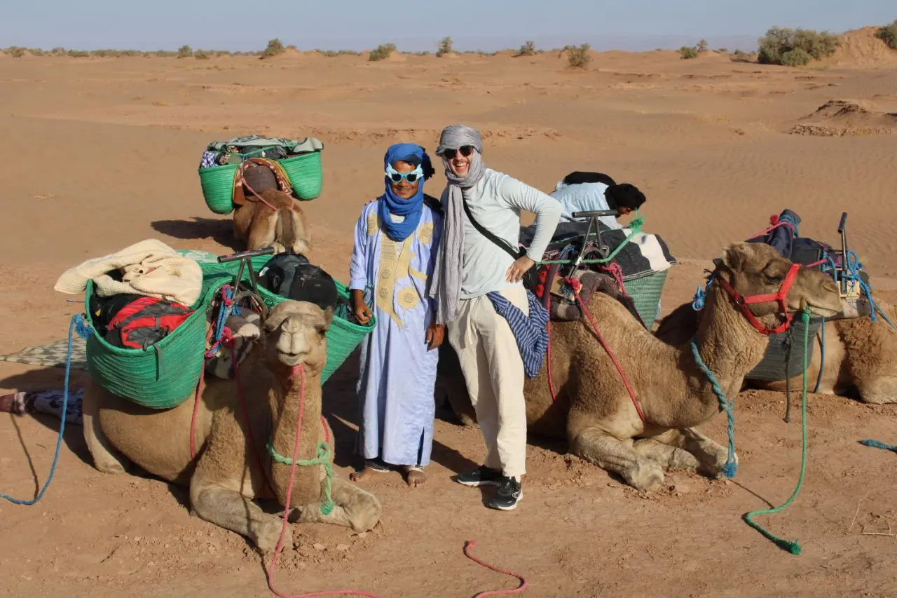 Two men, one in a blue robe, standing with loaded camels in a sandy desert.