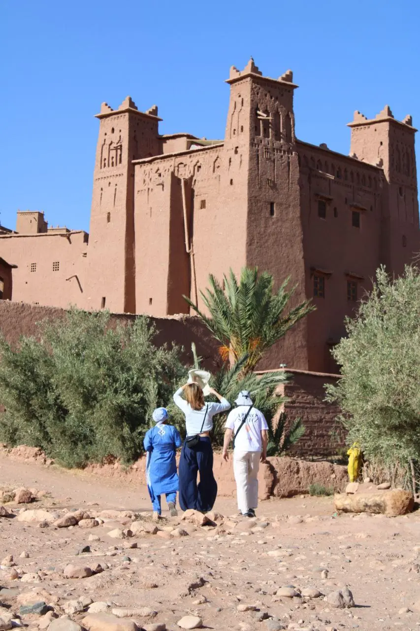 Three people walking towards the ancient mud-brick towers of a Moroccan kasbah under a clear blue sky.