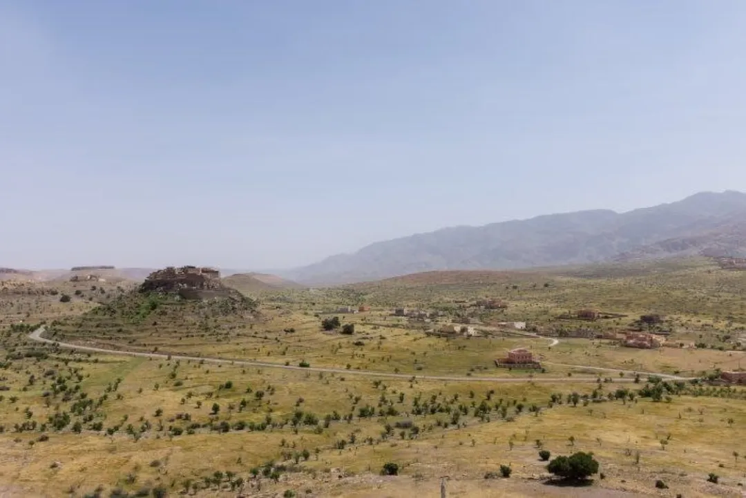 Arid valley with scattered trees, road, buildings, and distant mountains under a hazy sky