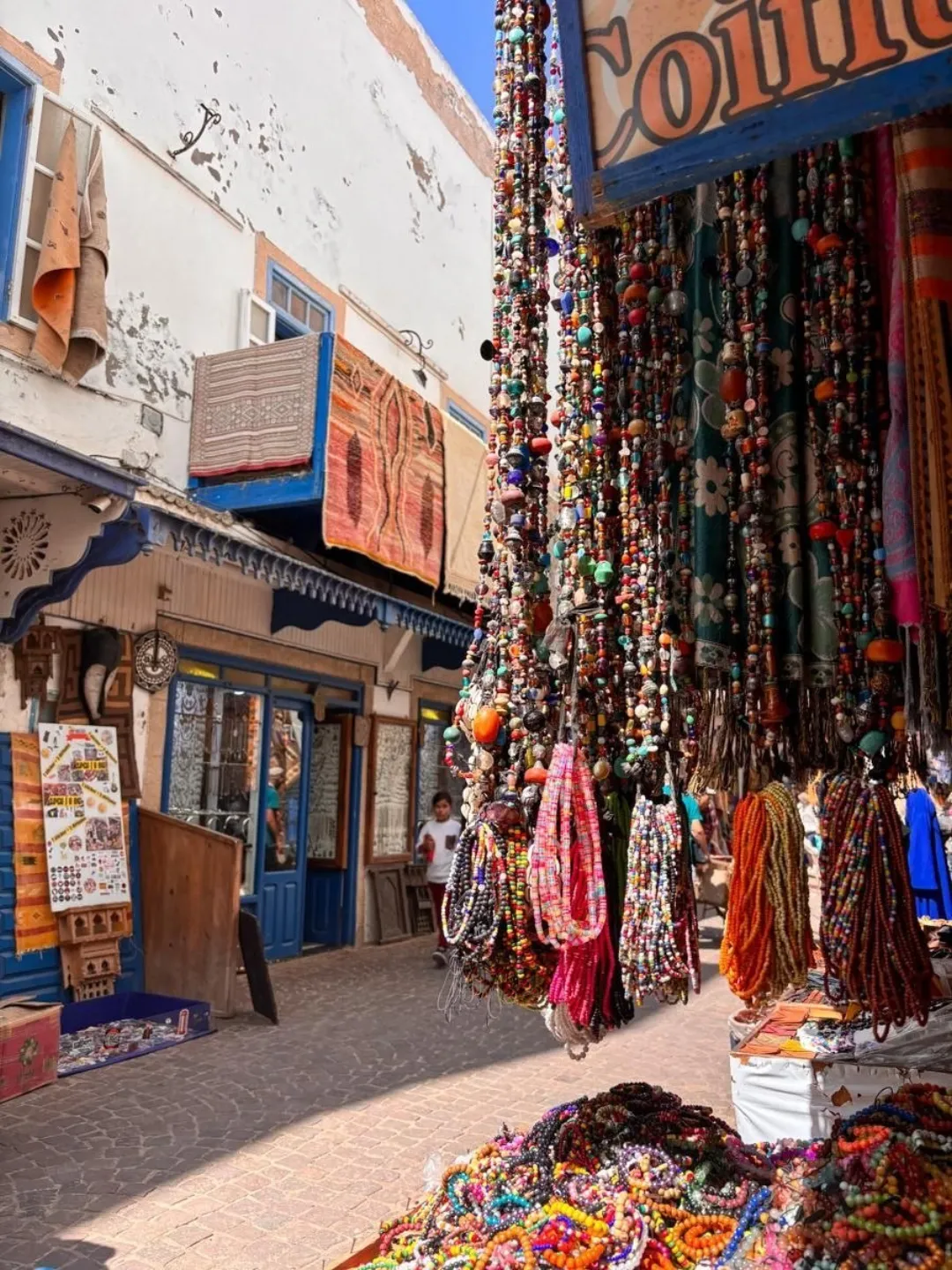 Colorful bead necklaces hanging in a narrow market alley with rugs and blue-trimmed shopfronts