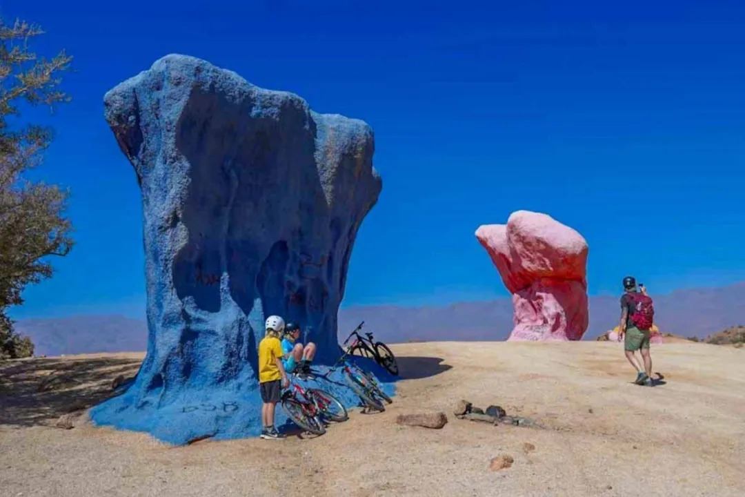 Cyclists beside large blue and pink painted rocks in a desert landscape
