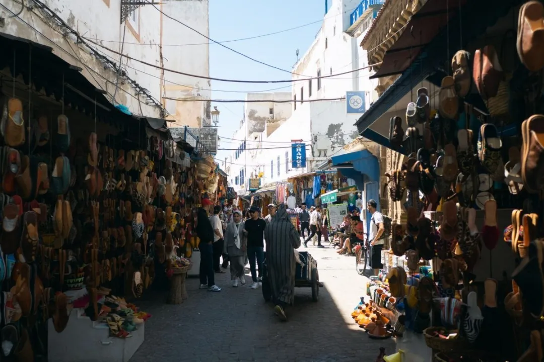 Shaded market alley lined with hanging leather slippers and shoppers