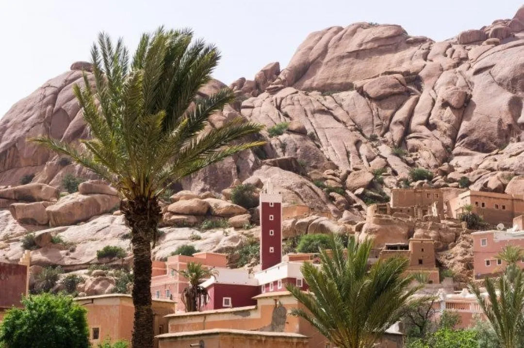 Palm trees and village buildings beneath large pink granite boulders in a desert oasis