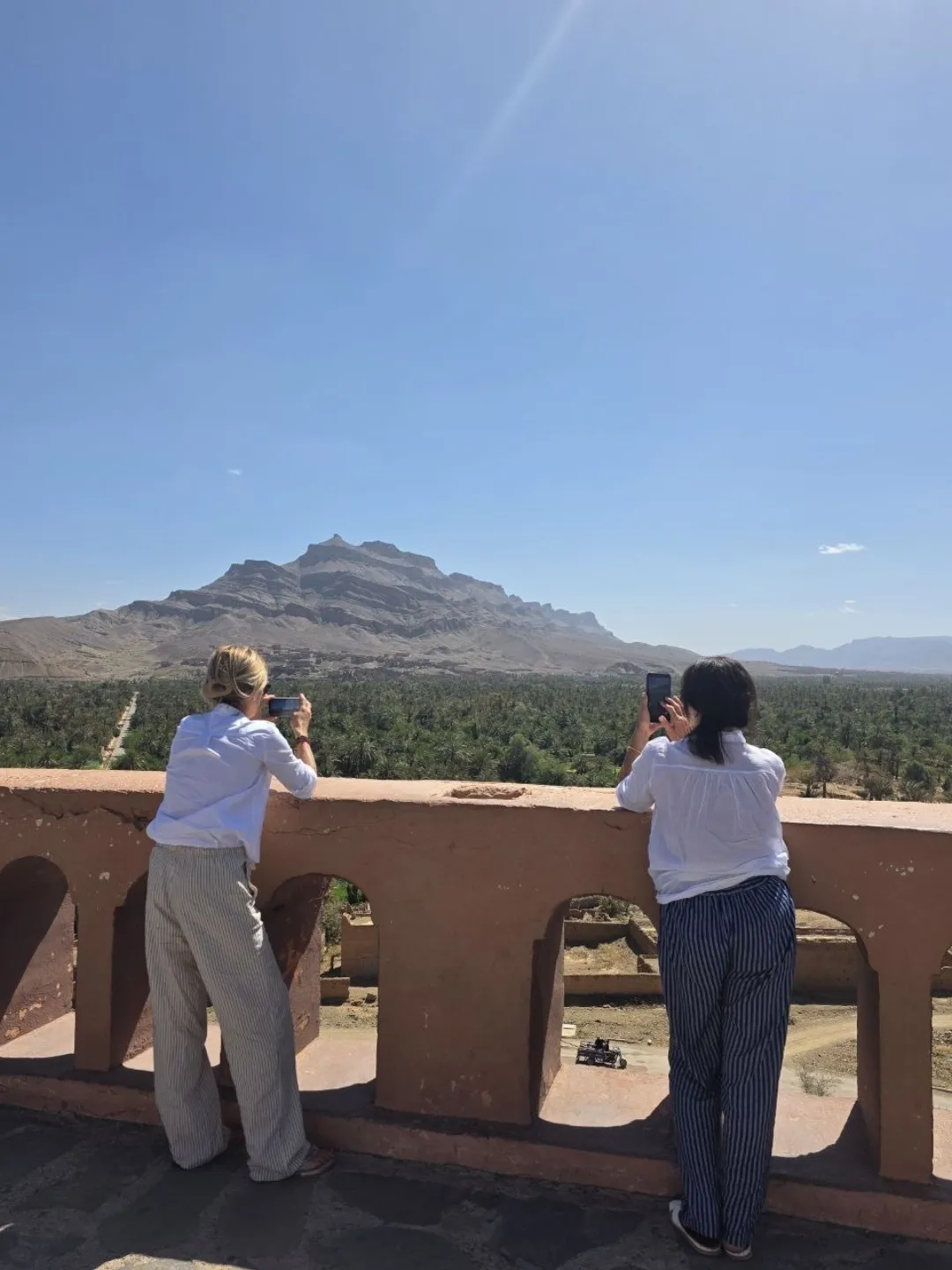 Two travelers photograph a palm oasis and rocky desert mountains from a terrace