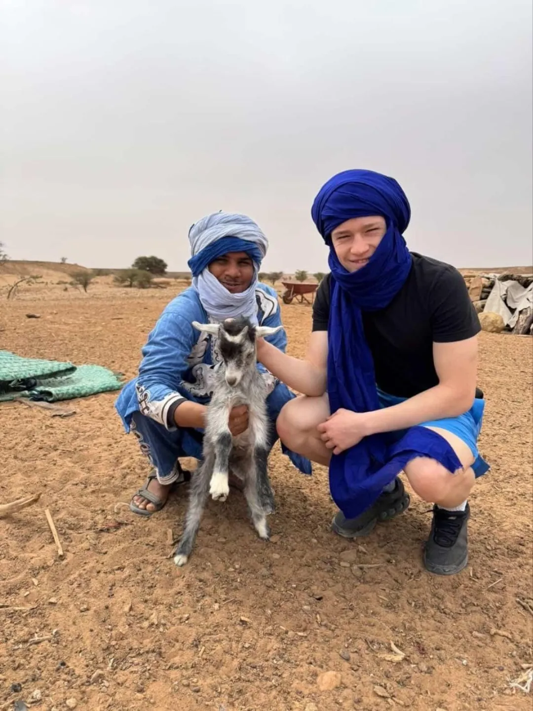 Two men in blue desert headwraps posing with a young goat on arid ground