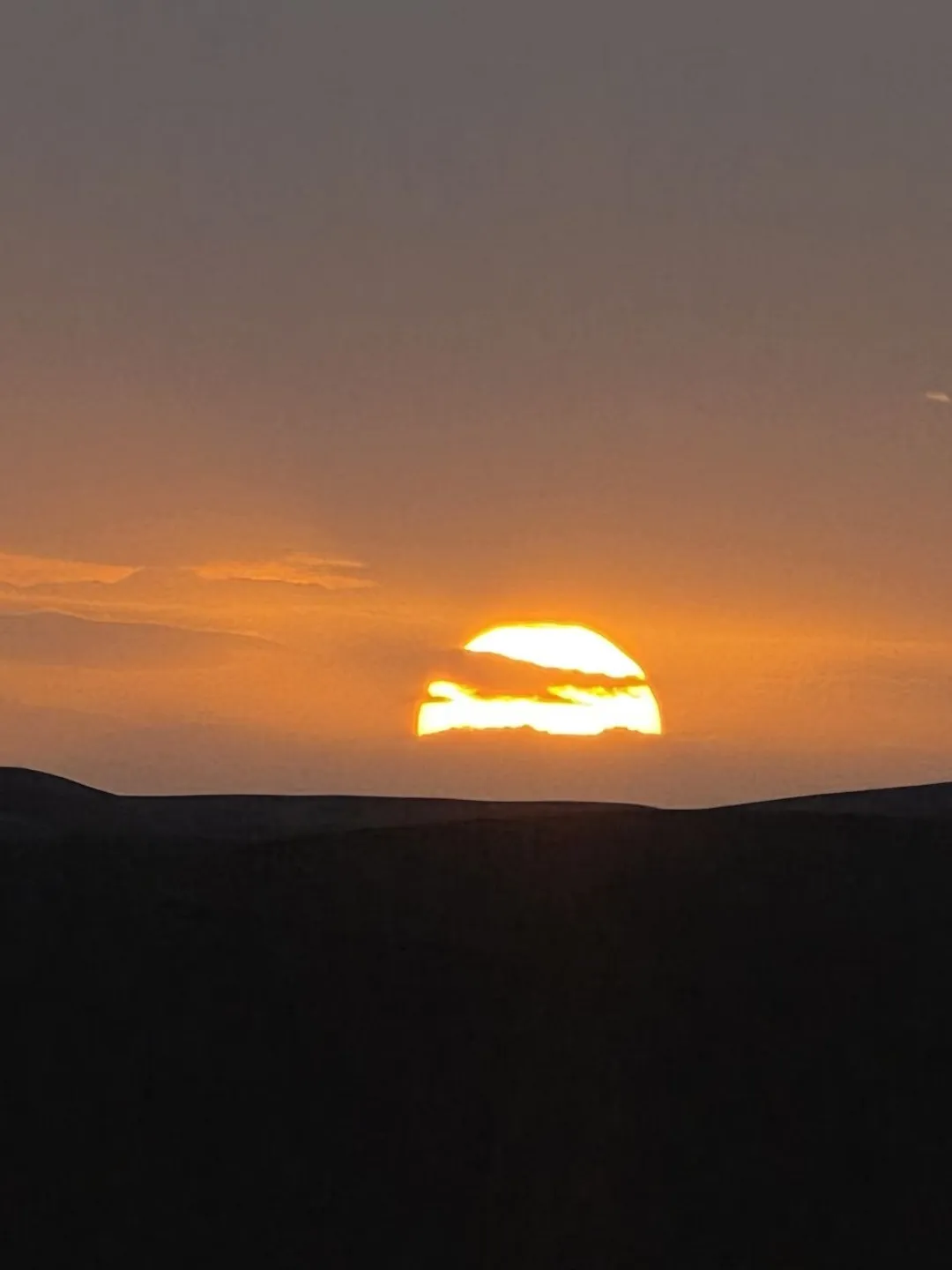 Sun setting behind dark desert dunes under an orange sky