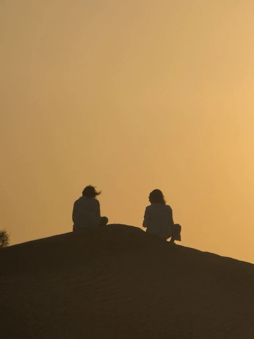 Two silhouetted people sitting on a sand dune at sunset