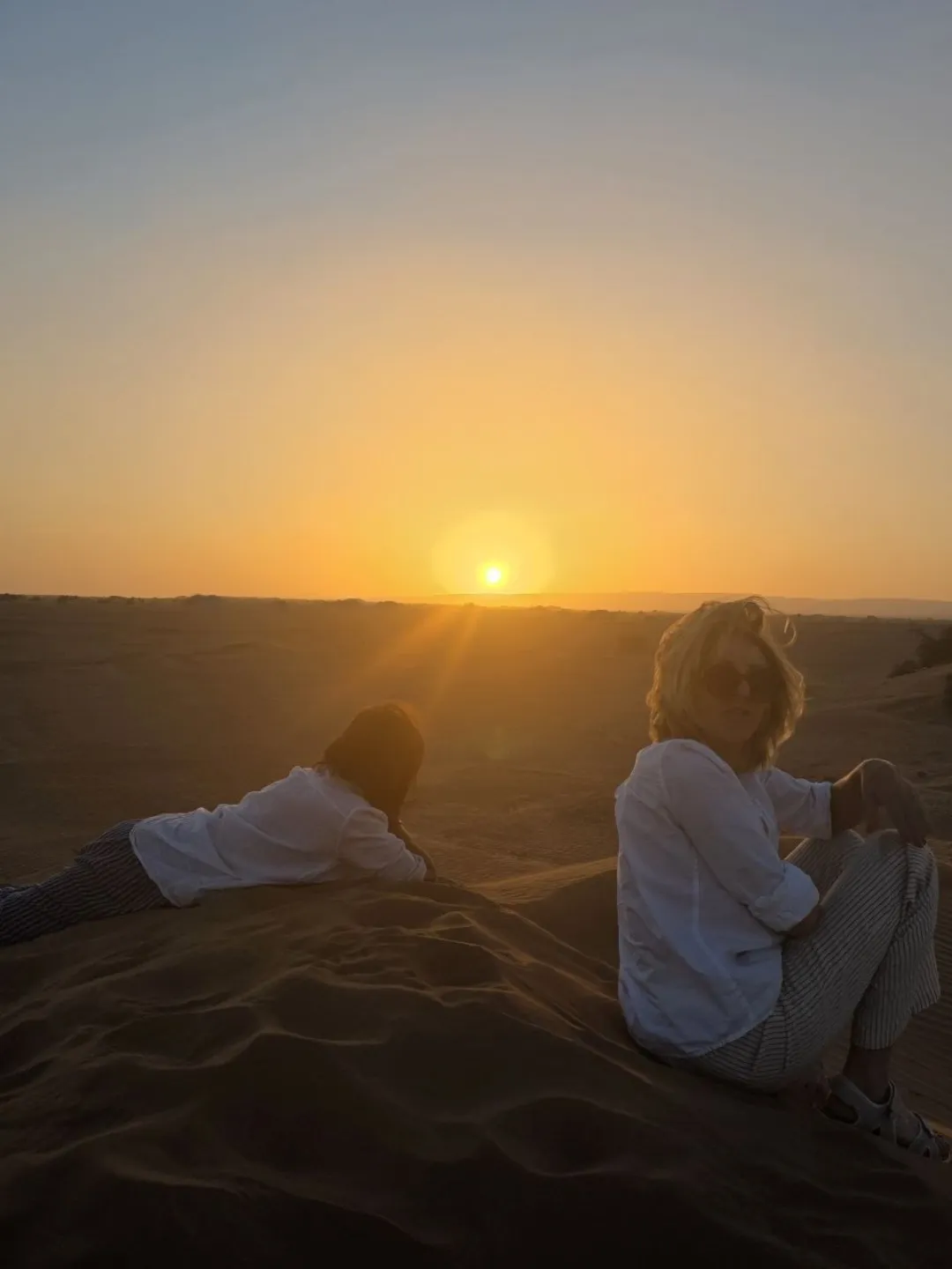 Two people on sand dunes watching a bright desert sunset.