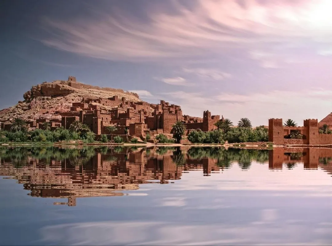 Desert kasbah village reflected in calm water beneath a pink sky
