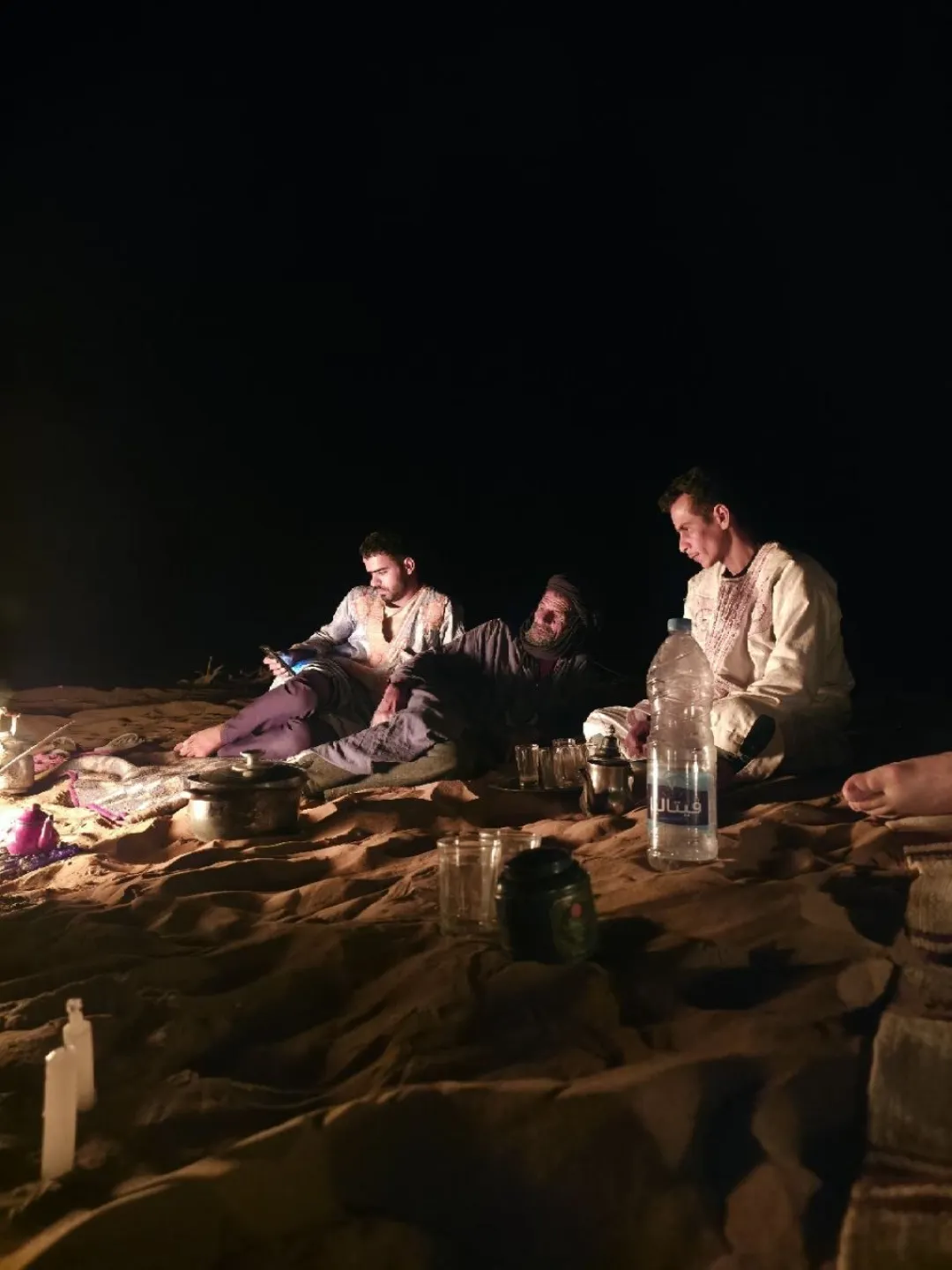 Three men sit on blankets in the Sahara at night with tea and a lantern